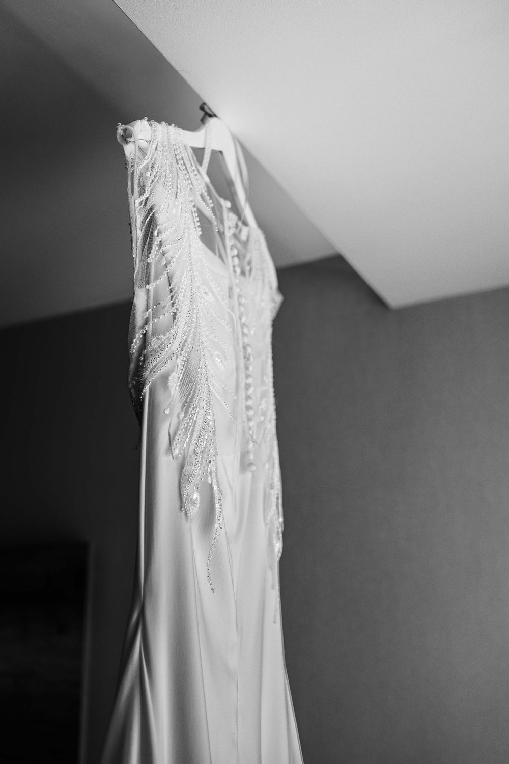 A delicate white wedding dress with intricate beading and lace details hangs against a wall.