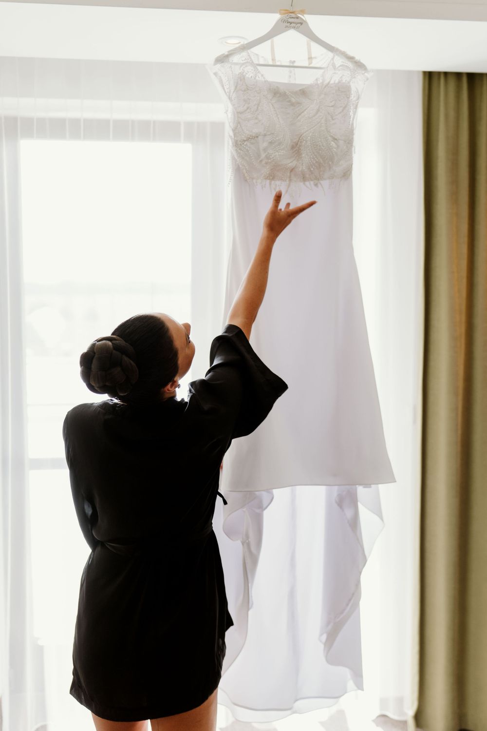Silhouette of person in black robe reaching up to touch white wedding dress hanging by window.