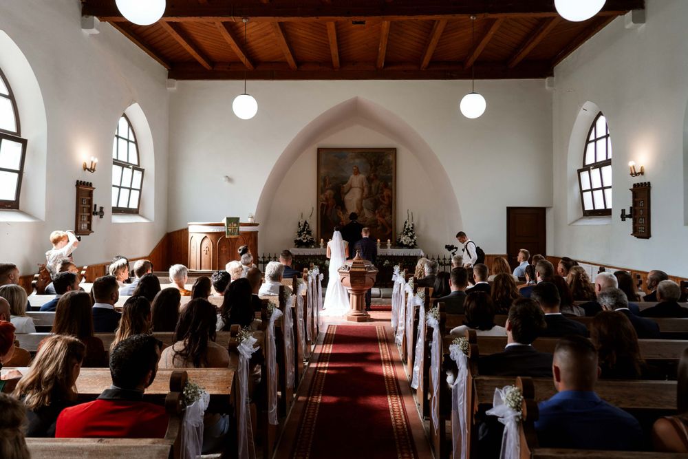 Church wedding ceremony with guests seated in wooden pews under arched windows and pendant lights.