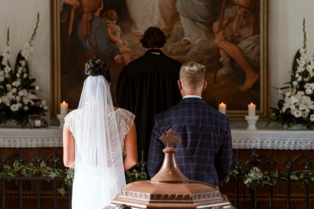Couple standing at altar during wedding ceremony with religious painting and floral decorations.