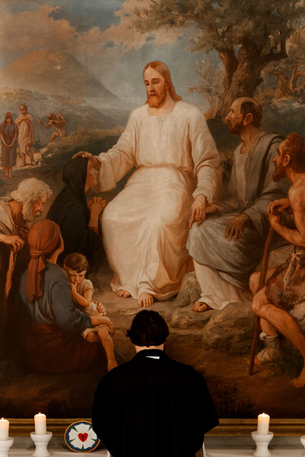 A religious painting depicting Jesus teaching while surrounded by listeners in a pastoral setting with lit candles below.
