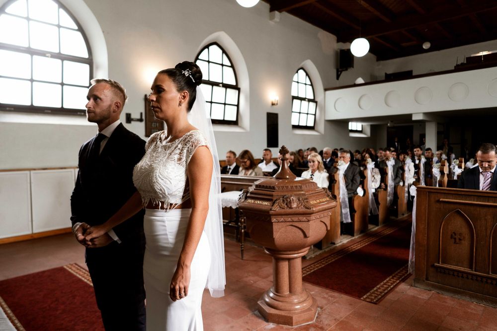 A couple stands at the front of a church with wooden pews and arched windows visible.