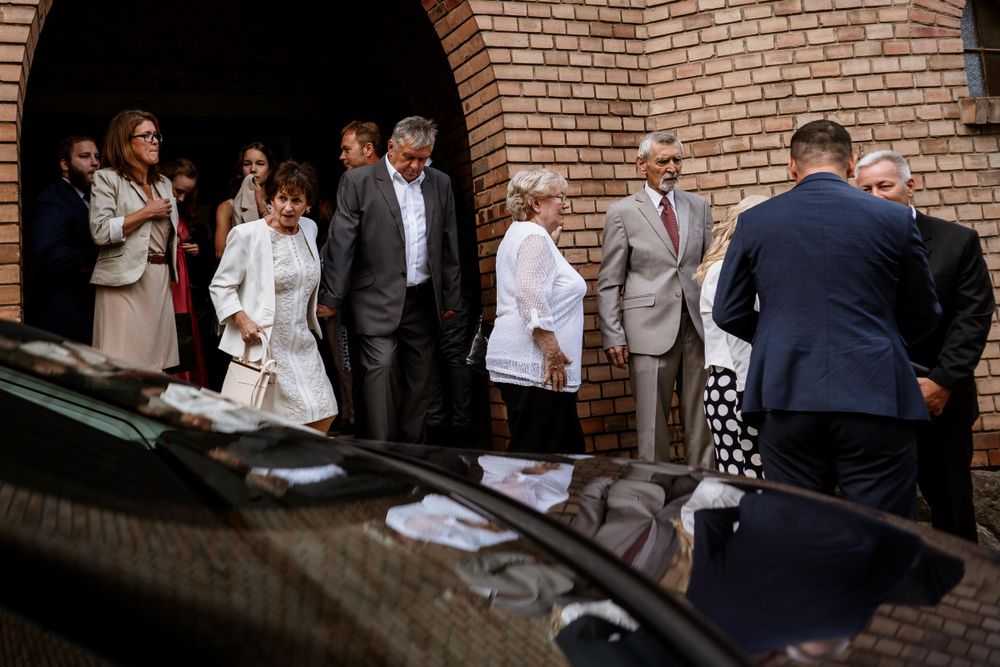 Wedding party gathered outside a brick church entrance near a parked car.
