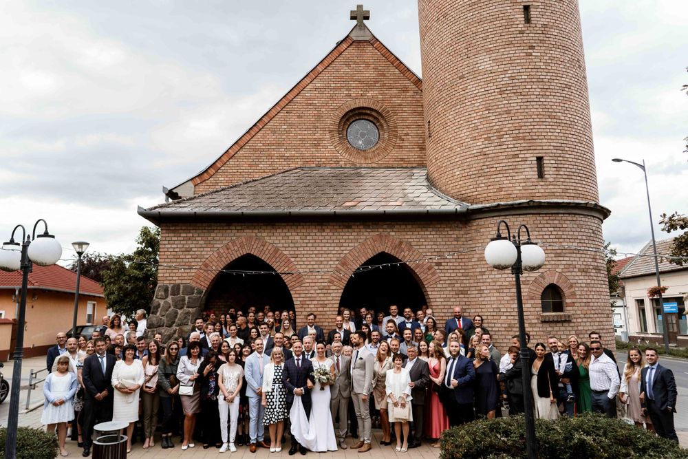 Large group wedding photo outside brick church with bell tower.