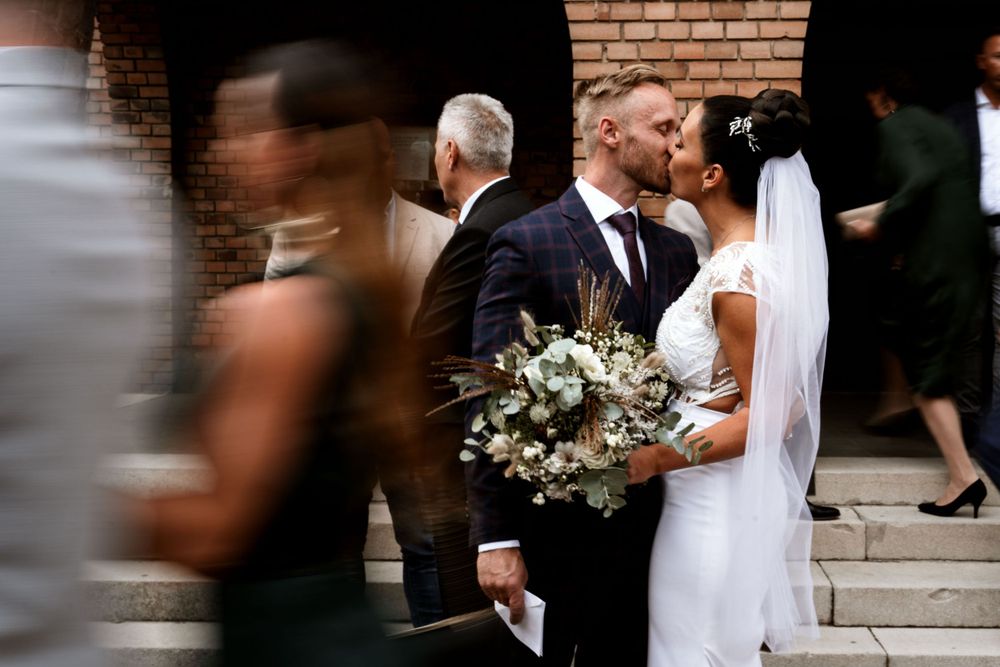 A couple with wedding bouquet embracing outside a brick building as guests move past.