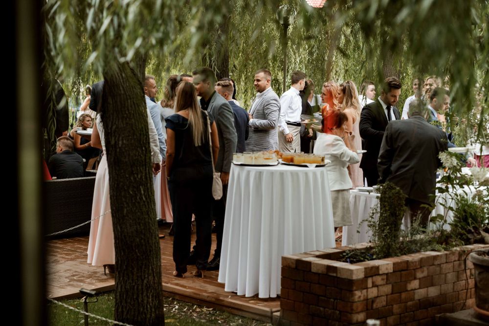 Wedding reception guests gather under willow trees near a white cocktail table.