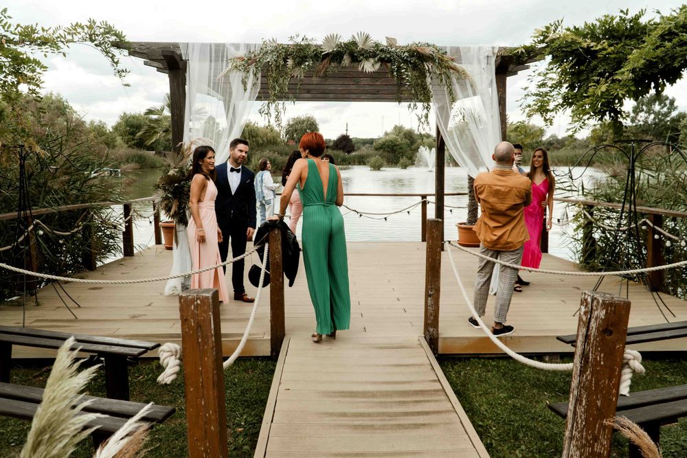Wedding guests gathering on wooden dock decorated with white fabric and floral arch.