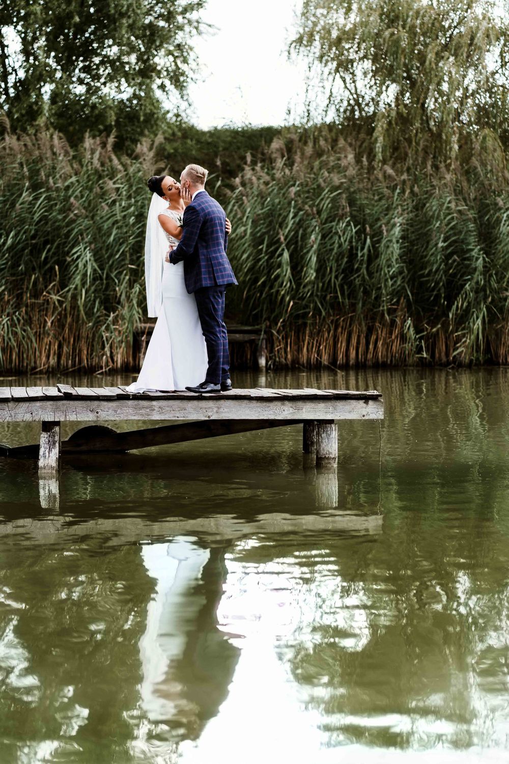 Romantic wedding couple stands on dock over reflective pond with willow trees in background.