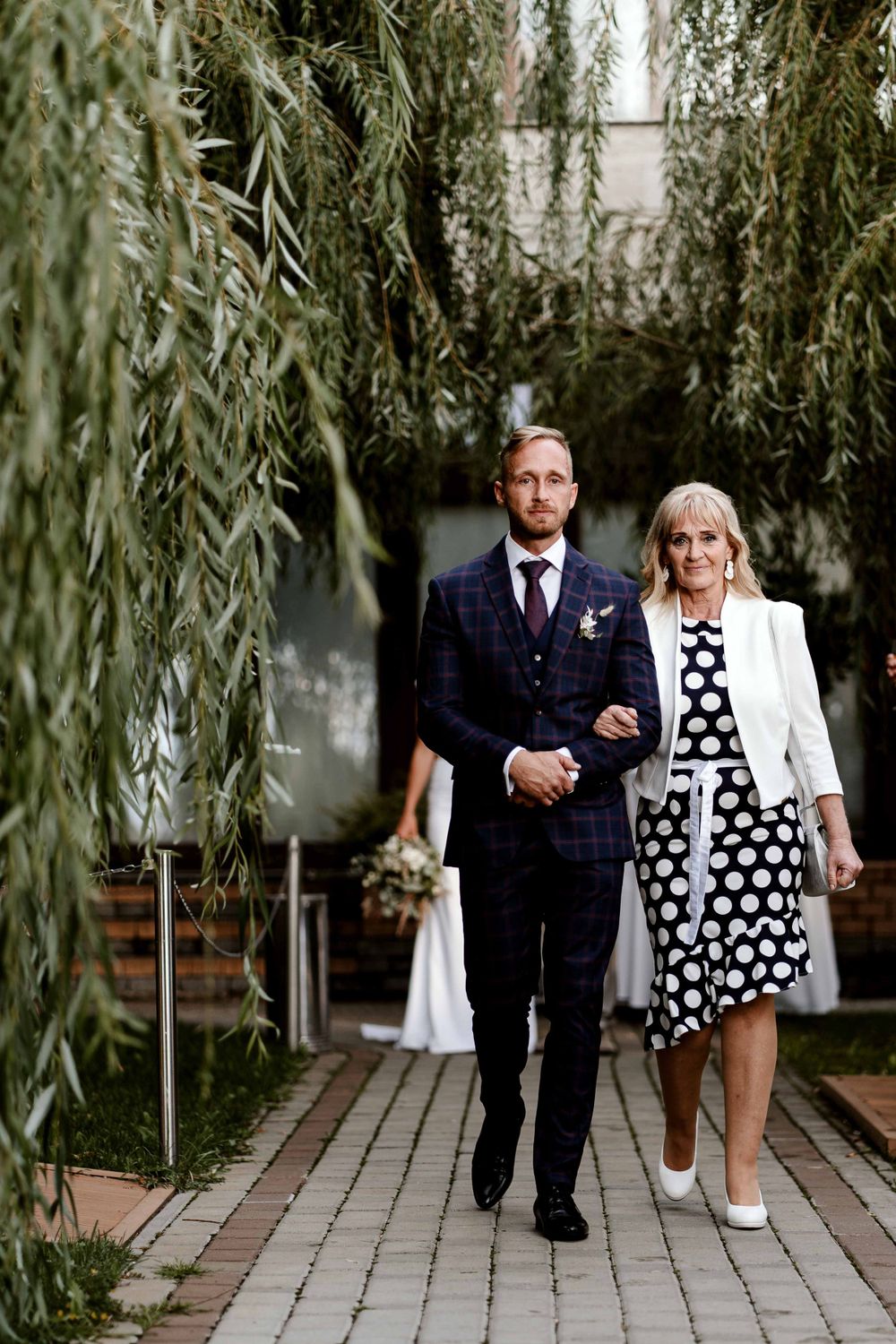 A couple walking under willow trees along a stone path at a garden venue.