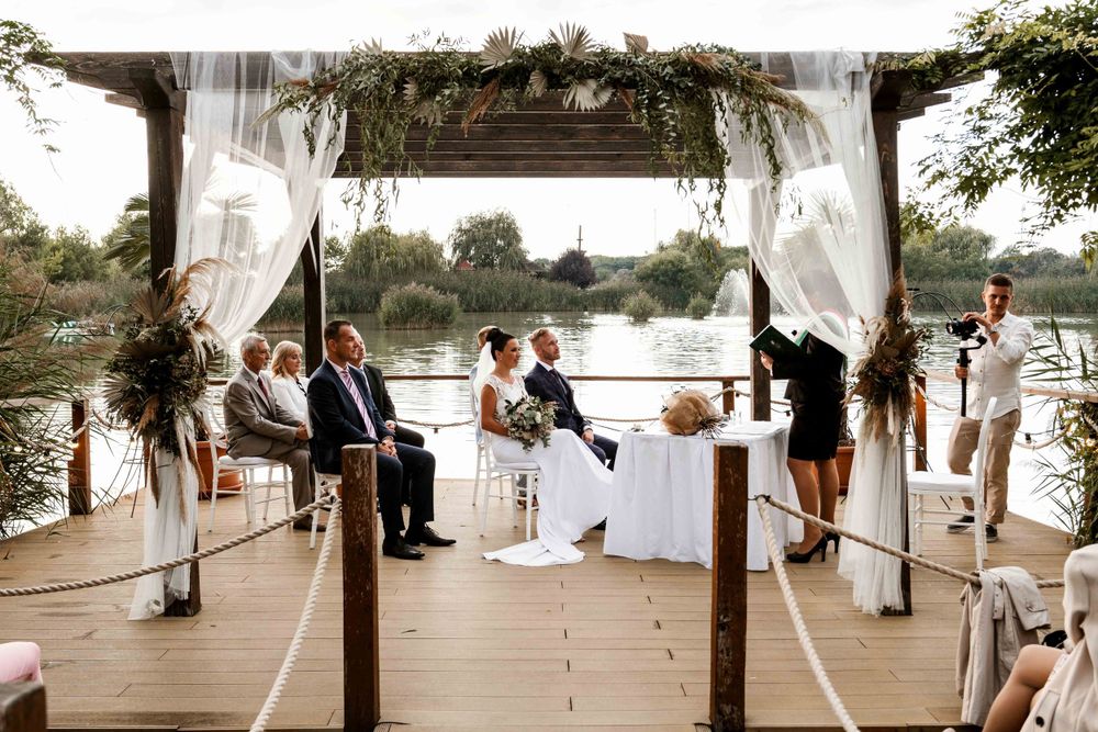 Outdoor wedding ceremony under a pergola decorated with white drapes and greenery by the waterfront.