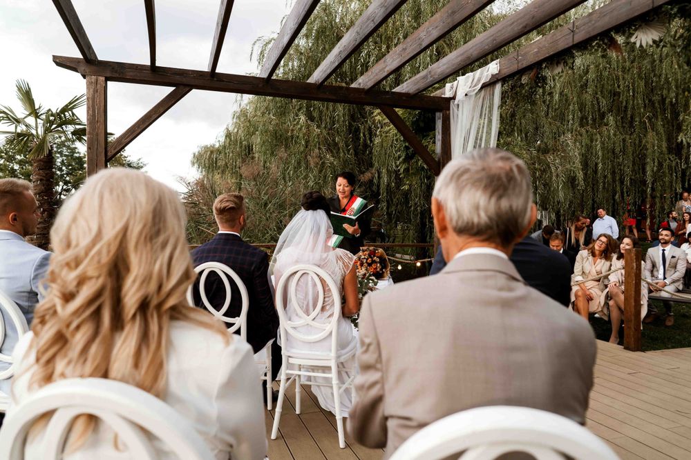 Wedding guests seated in white chairs watching ceremony under a draped pergola.