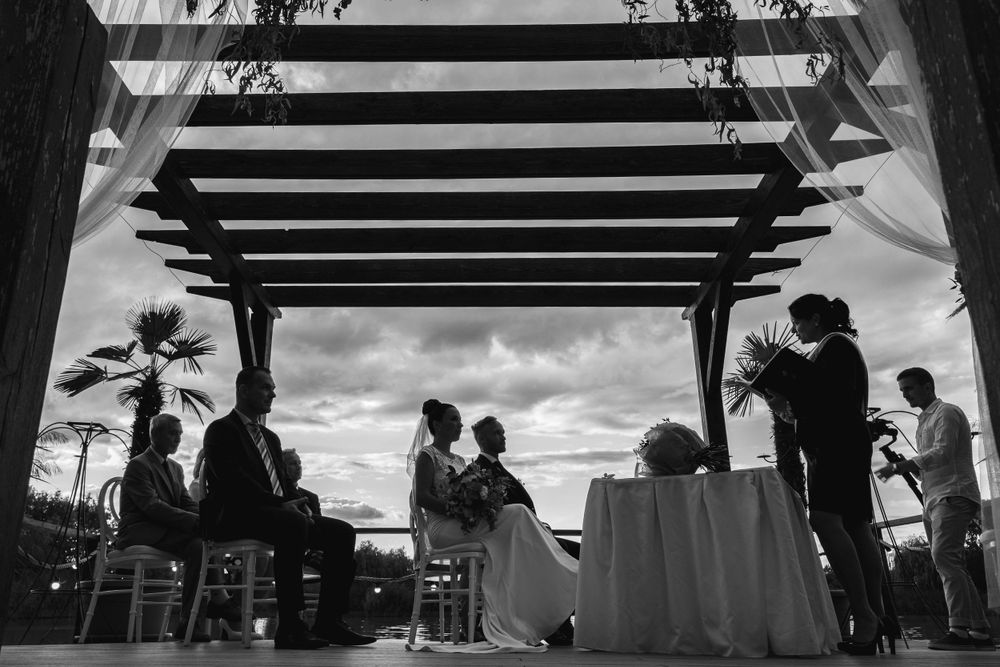 Silhouetted wedding ceremony under wooden pergola at sunset with guests seated around decorated table.
