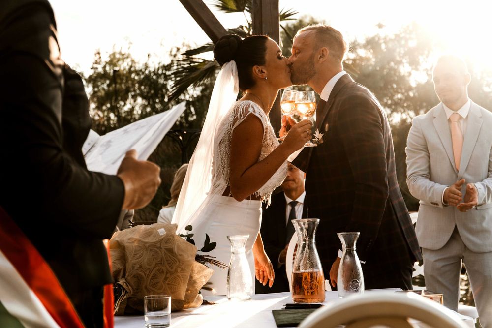 Sunlit wedding ceremony under wooden pergola with orange drinks on table.