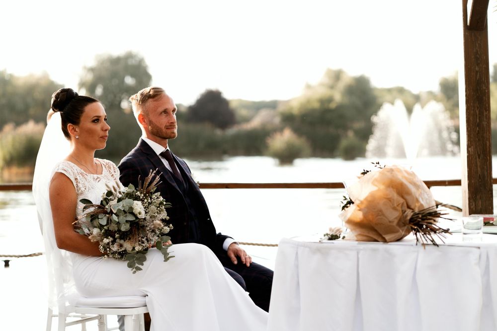 A bride and groom sit together during their outdoor wedding ceremony beside a fountain.