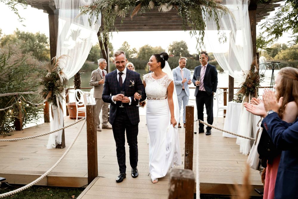A couple walking through a decorated outdoor wedding arch with white draping and greenery.