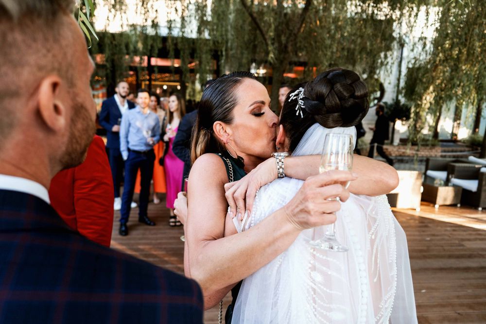 Wedding guests embrace and share celebratory drinks on an outdoor deck beneath willow trees.