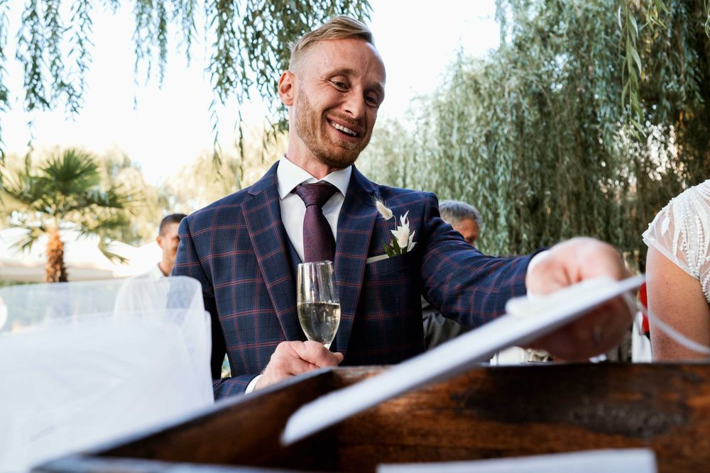 Guest in plaid suit jacket holds champagne while smiling at outdoor wedding reception.
