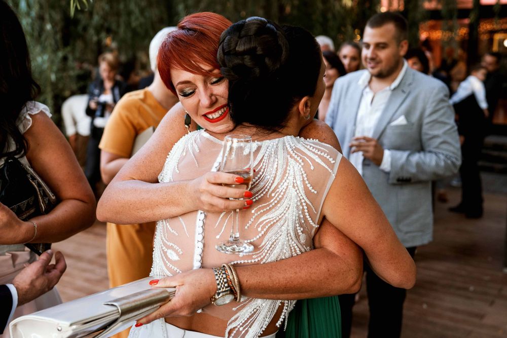 Wedding reception guests share warm hugs while holding champagne glasses outdoors.