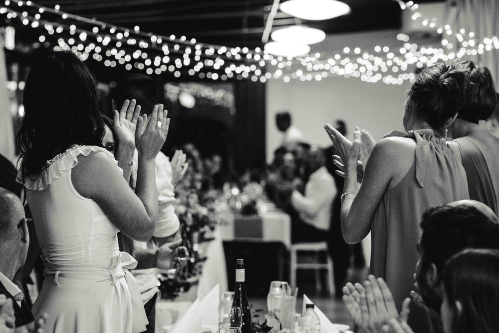 Wedding guests clap and celebrate during reception festivities under twinkling lights.