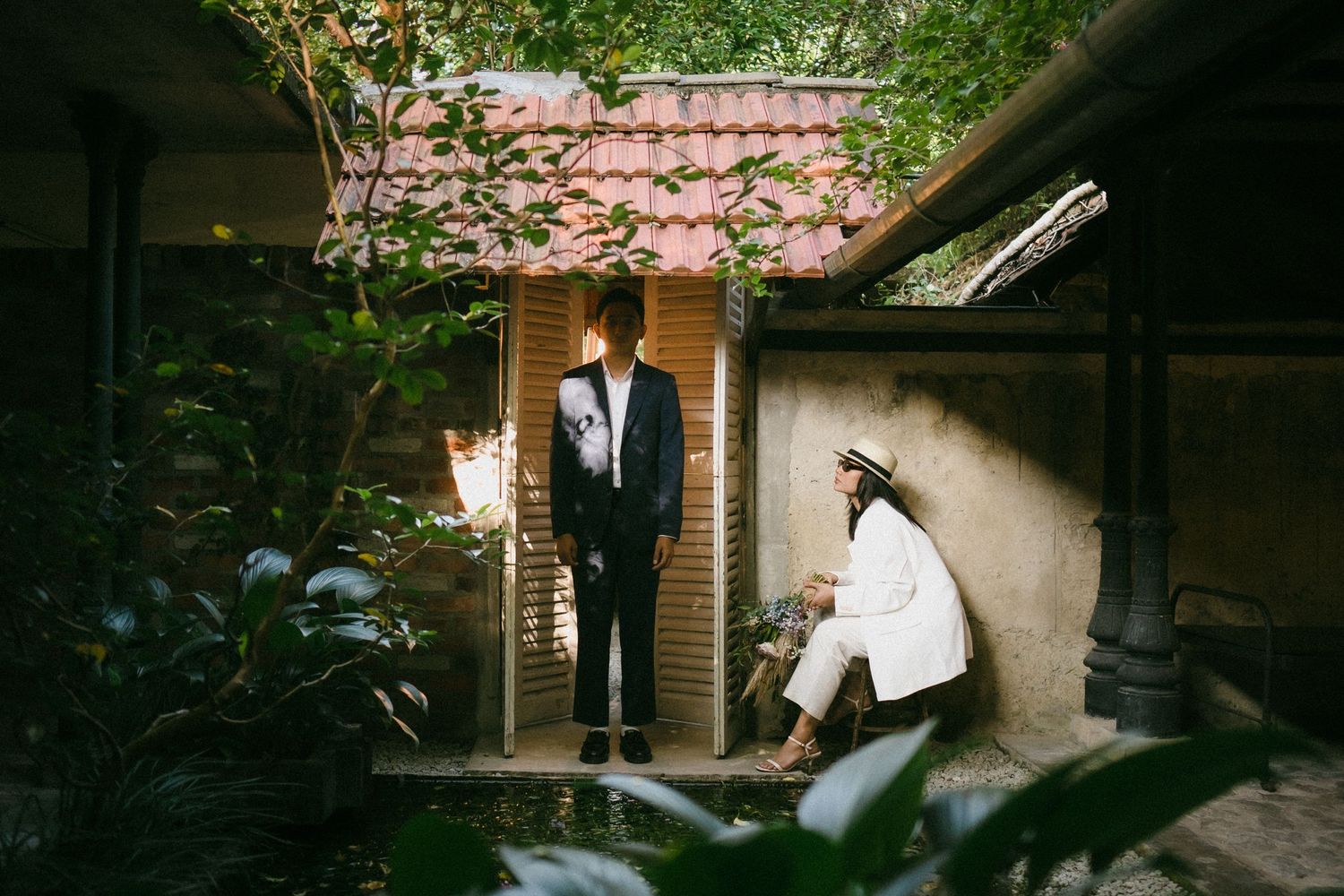 Wedding couple in a charming rustic courtyard surrounded by greenery, groom standing as bride sits nearby.