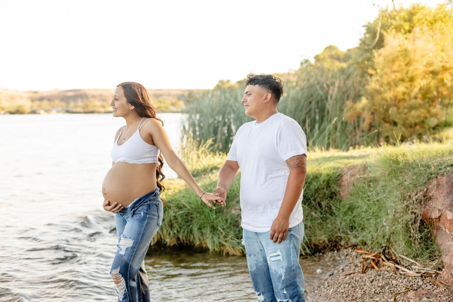 Expectant couple sharing a tender moment by a lake during sunset.