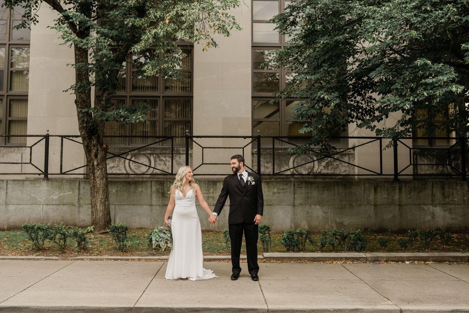 A couple in wedding attire poses together on a city sidewalk.