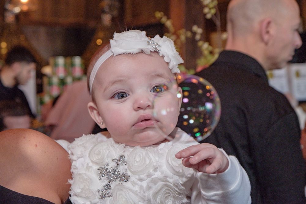 Baby wearing white dress and floral headband at formal event with soft bokeh lights in background.