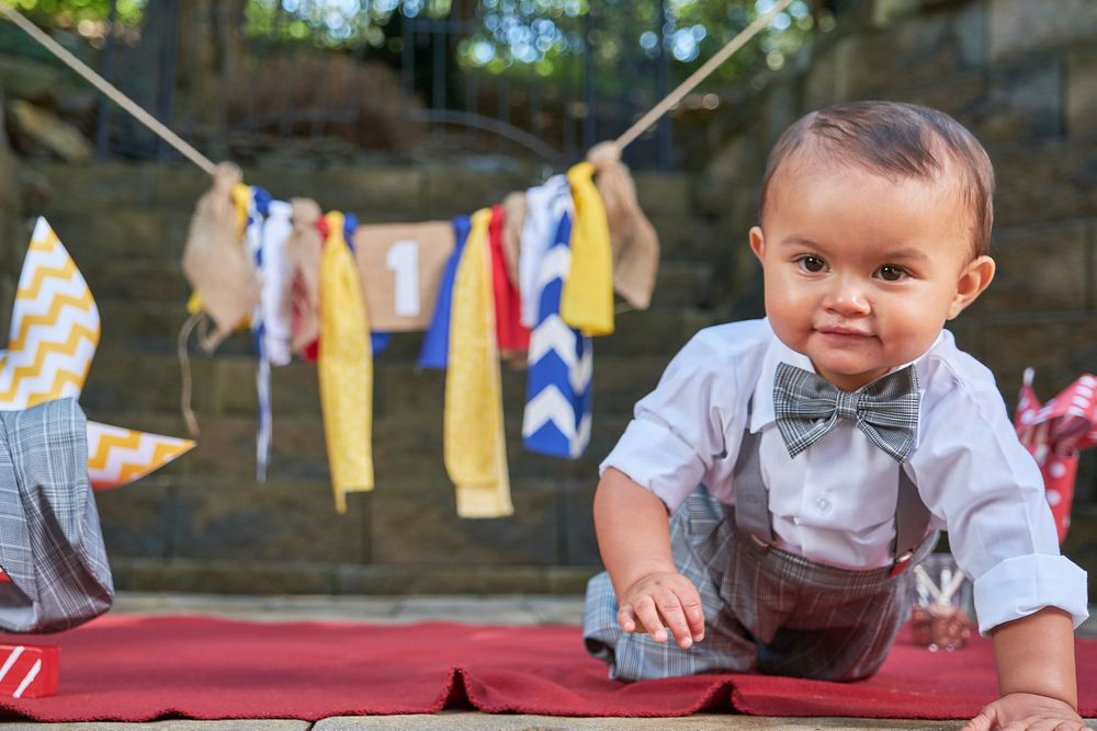 A baby in suspenders and bow tie crawls on red blanket with colorful bunting in background.