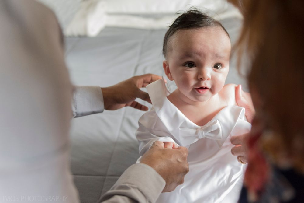 Baby in white clothing sitting on bed surrounded by people.