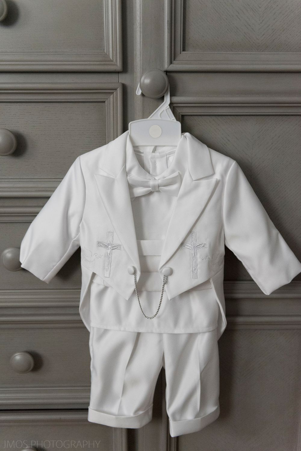 White baptismal outfit with tails and bow tie hanging against grey cabinet.