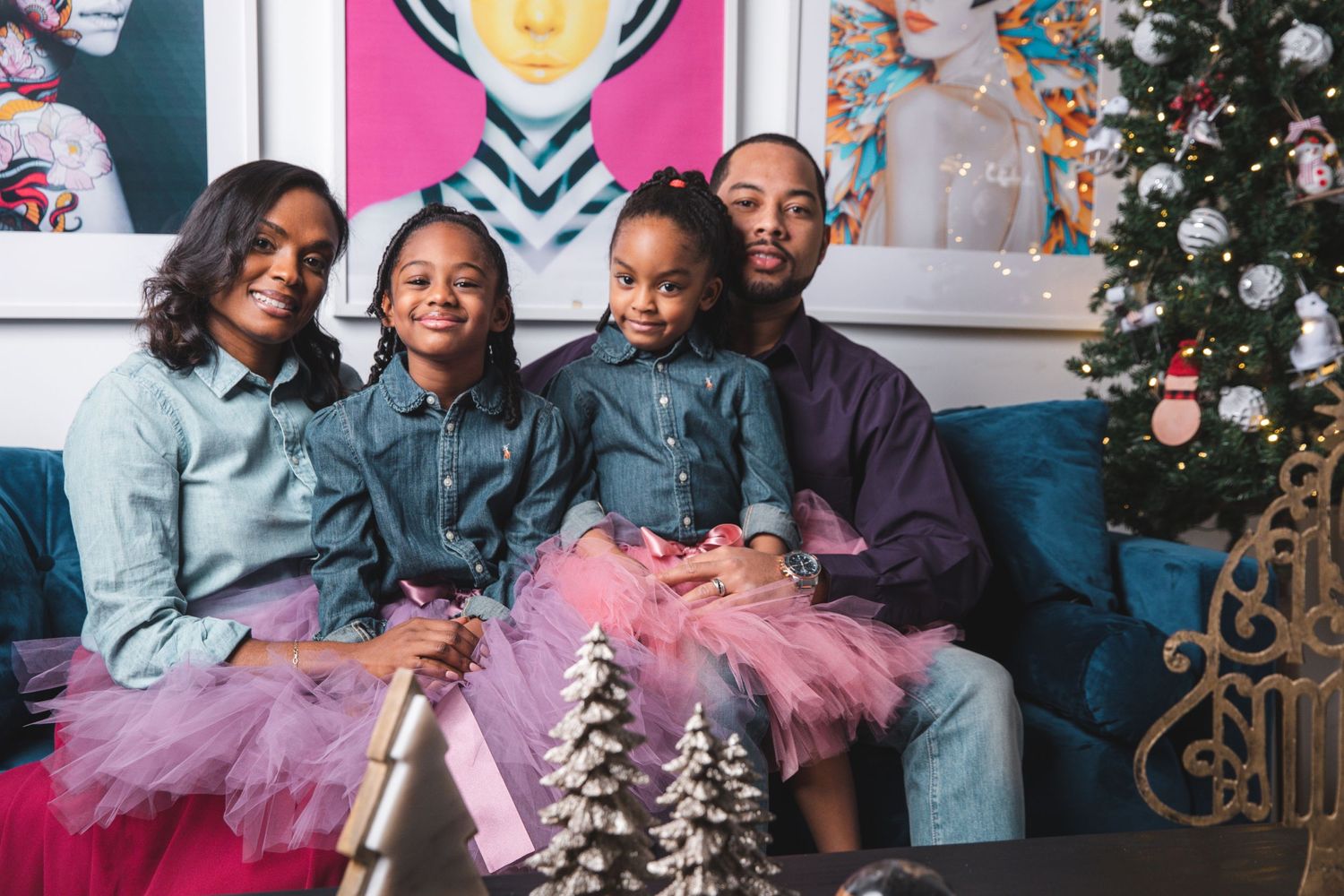 Black family of four in matching denim and pink tutu outfits smiling together near Christmas tree holiday decor.