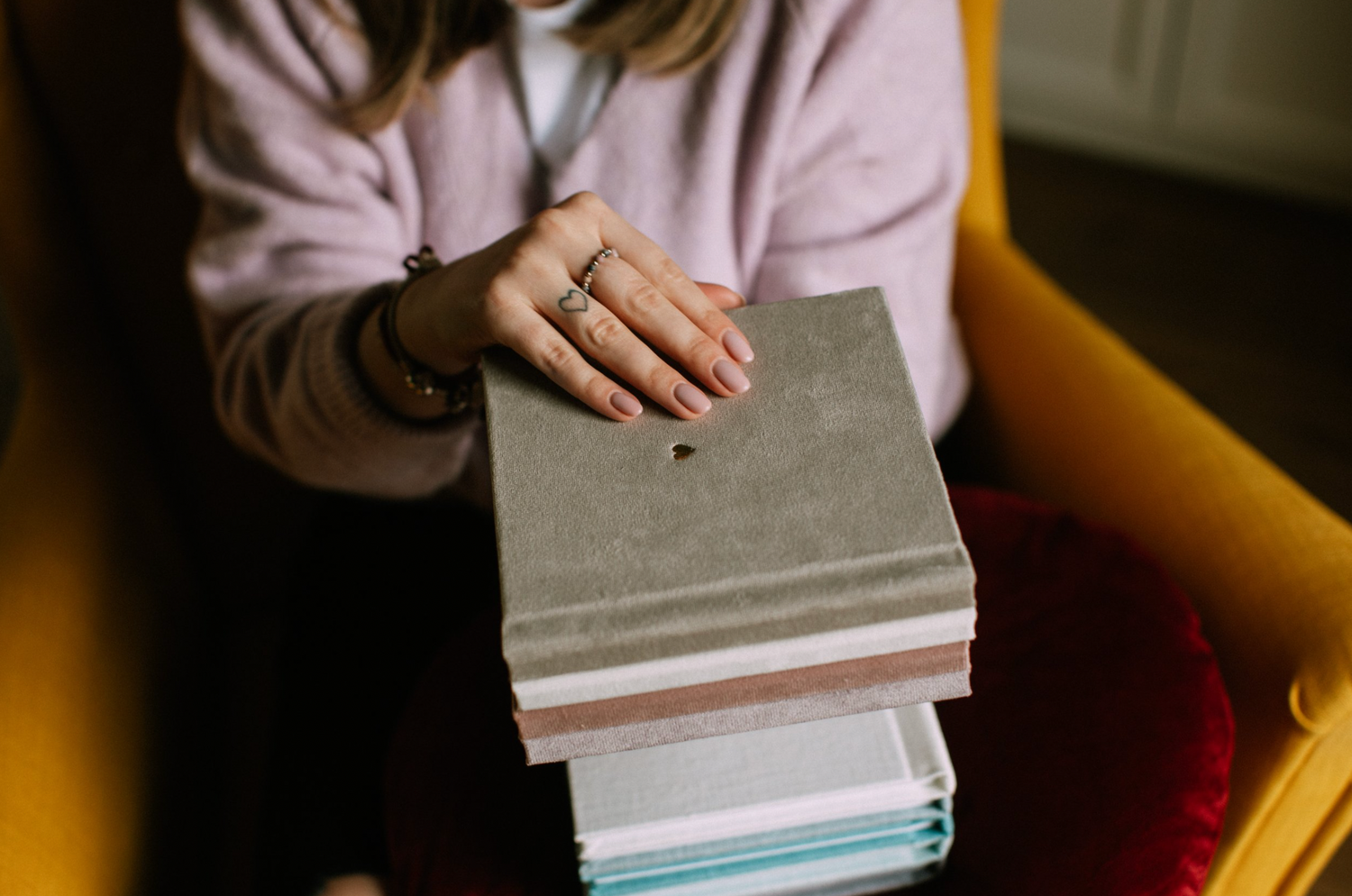 Hands holding a stack of photo albums in pastel colors while seated in a yellow chair.