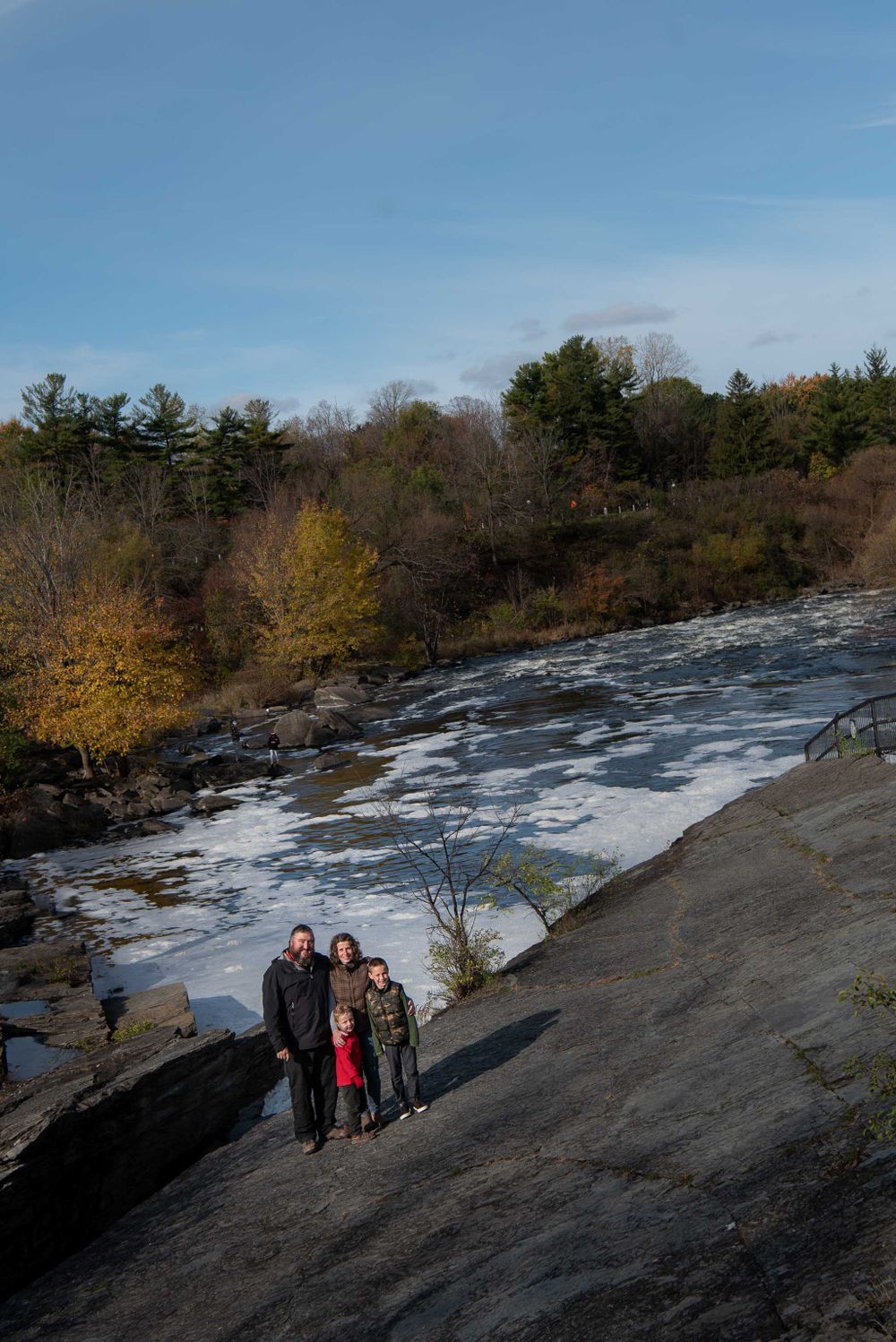 Colour family portrait taken from a distance and showcasing the small rapids and fall foliage in the forest behind them.
