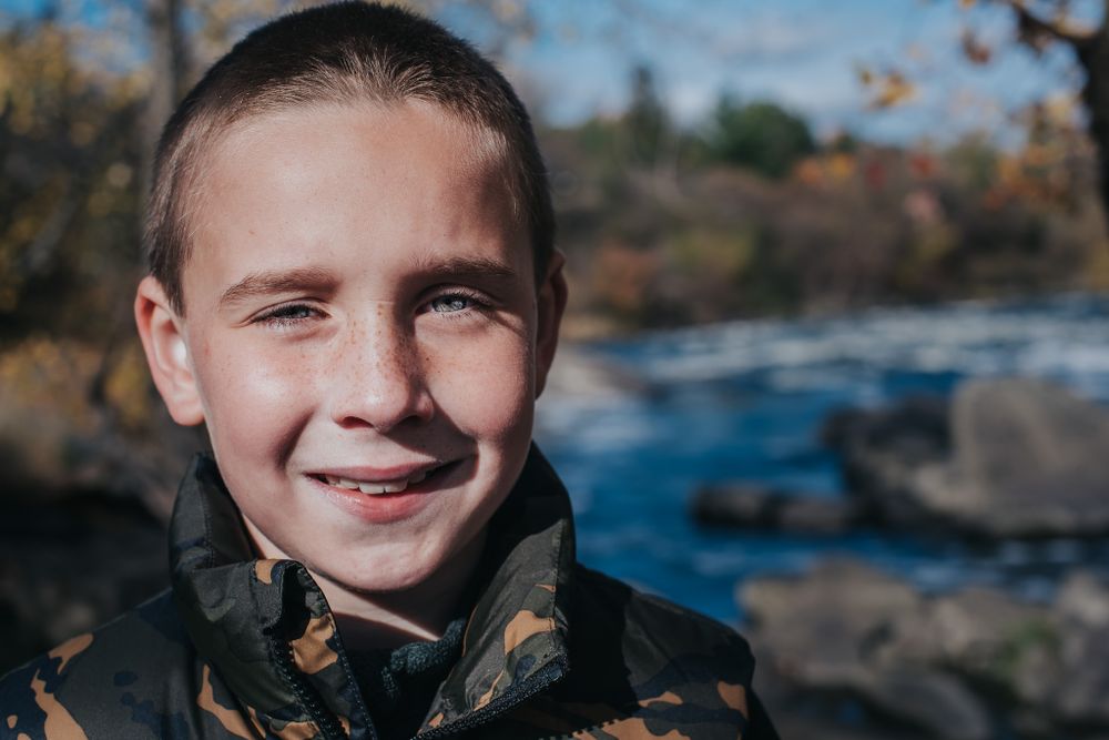 Colour portrait of a Caucasian boy smiling at the camera with water and fall forest in the background.