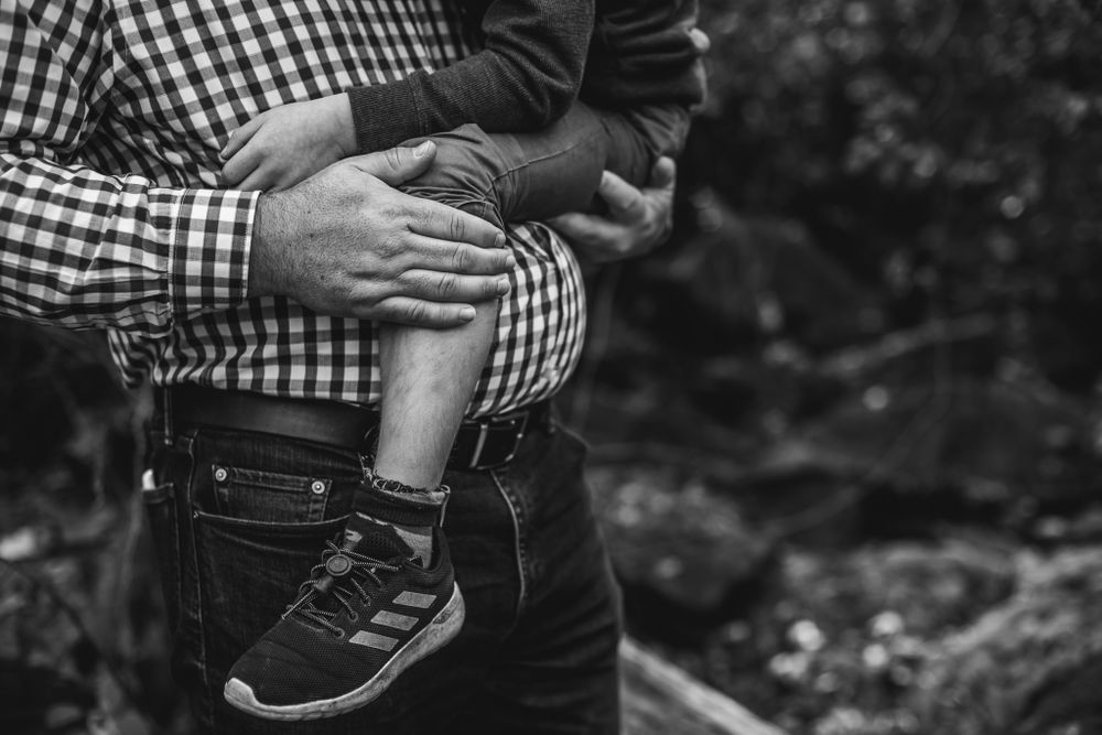 Image focused on a fathers hand as he holds his son in his arms and pats his knee after a fall.