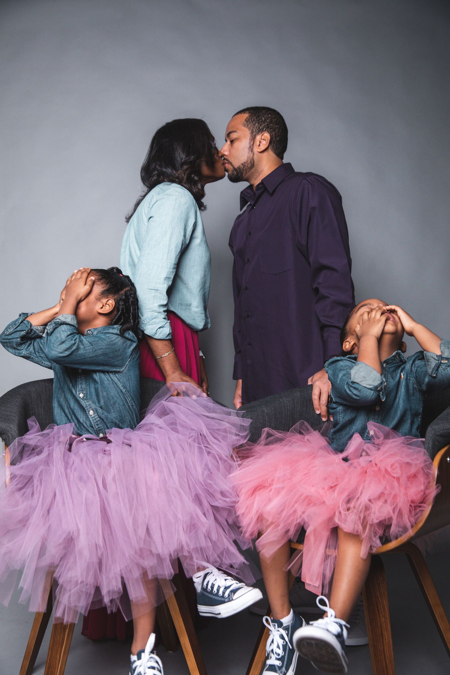 Black couple sharing a kiss while their two daughters in tutus cover their eyes during family photo session.