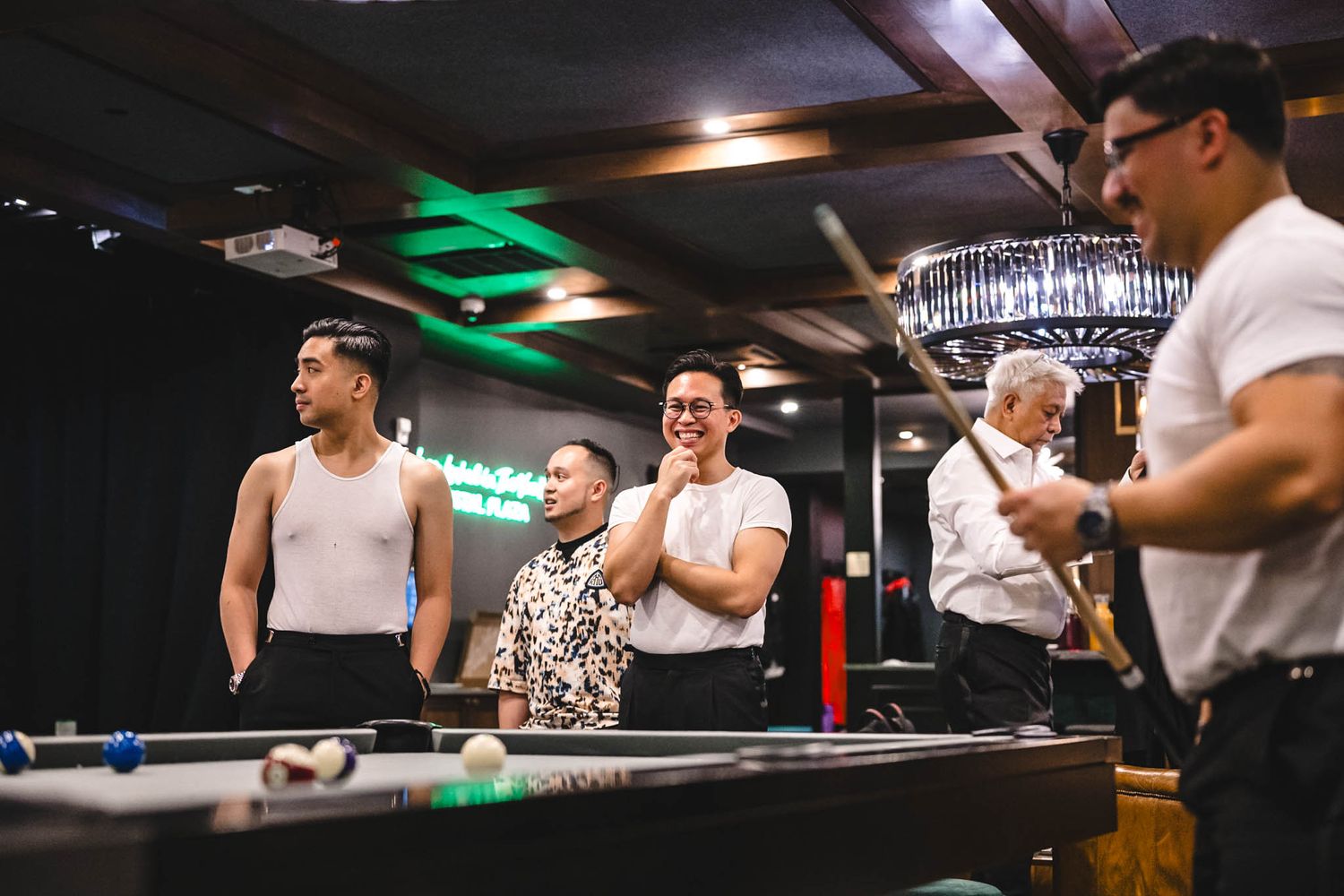 Groomsmen gather around a pool table in a dimly lit room with green lighting during a wedding celebration.