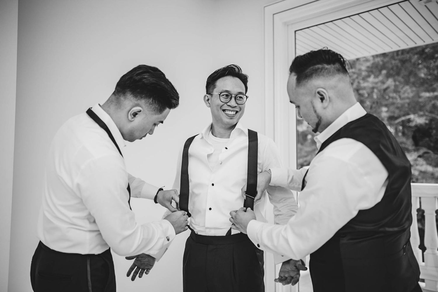 Three men in formal attire help each other get dressed before a wedding ceremony.