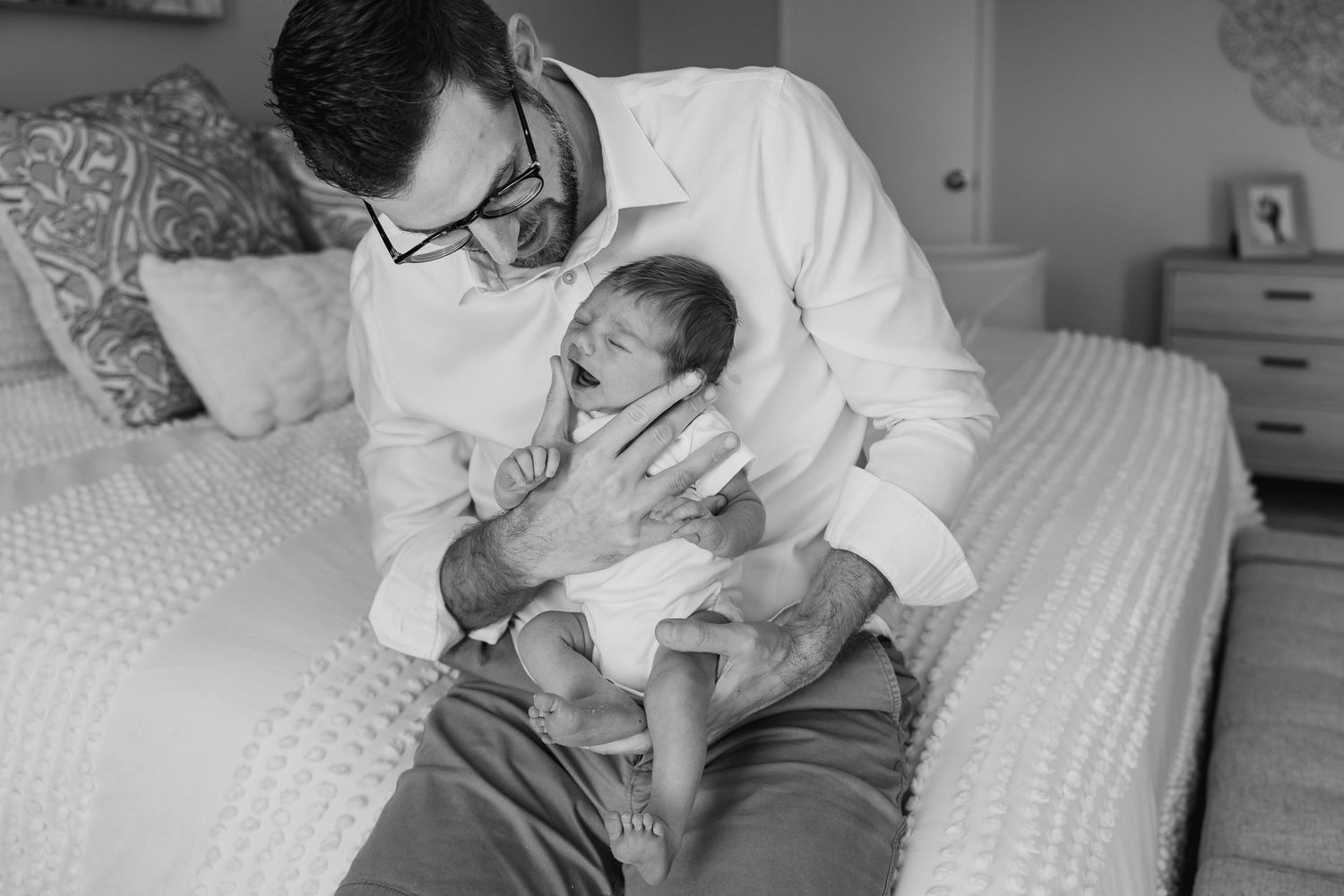 A tender black and white portrait of father and newborn baby resting together on a bed.