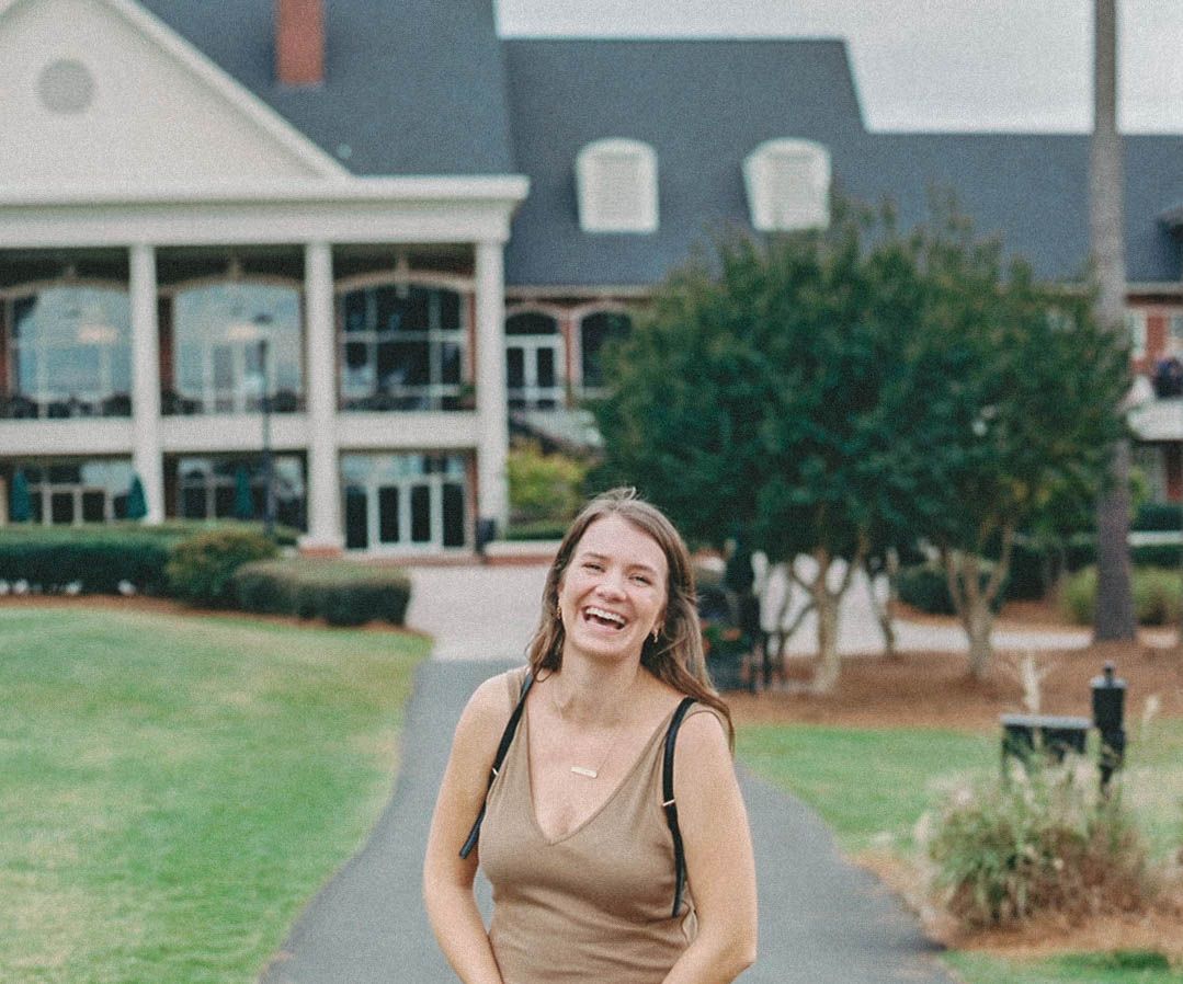 Happy young woman laughing in front of a grand white colonial-style building on green lawn.
