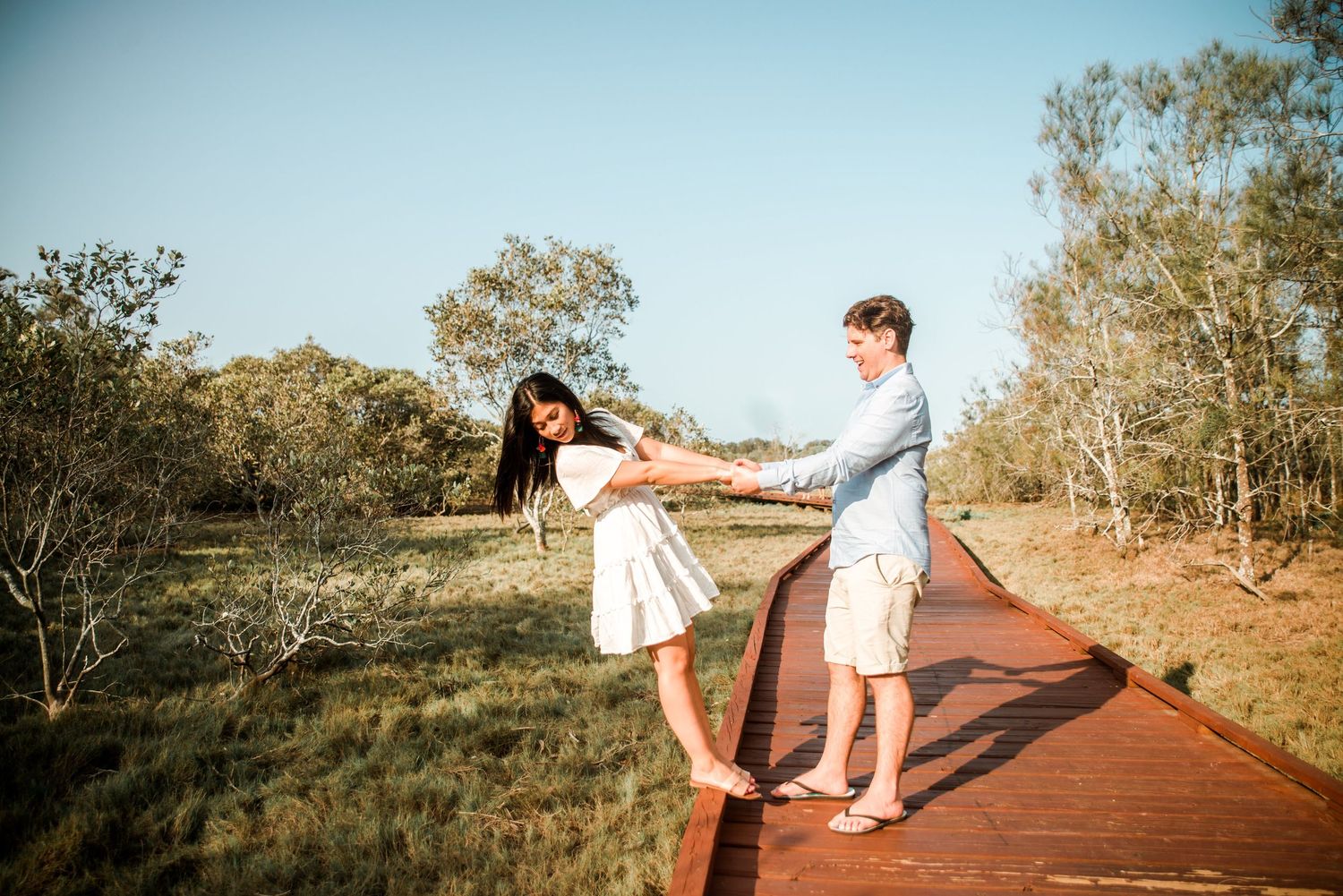 A playful couple dances and spins on a wooden boardwalk surrounded by coastal vegetation.