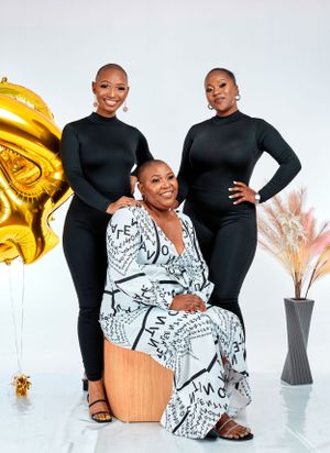 Three women in coordinated outfits pose together with a gold balloon against a light background.
