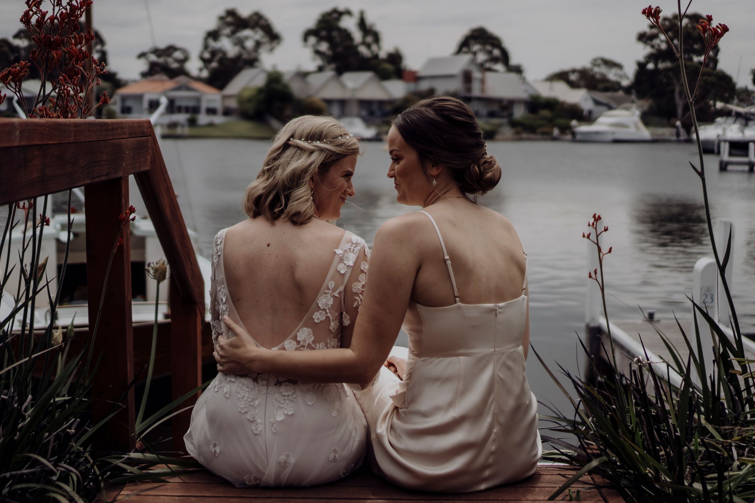 Two people in elegant dresses sit close together on a wooden dock by a tranquil lake at dusk.