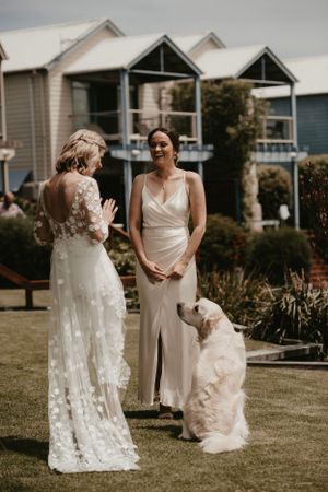 Two brides in white dresses share a joyful moment on a lawn with a fluffy dog while houses stand in the background. Paynesville wedding.