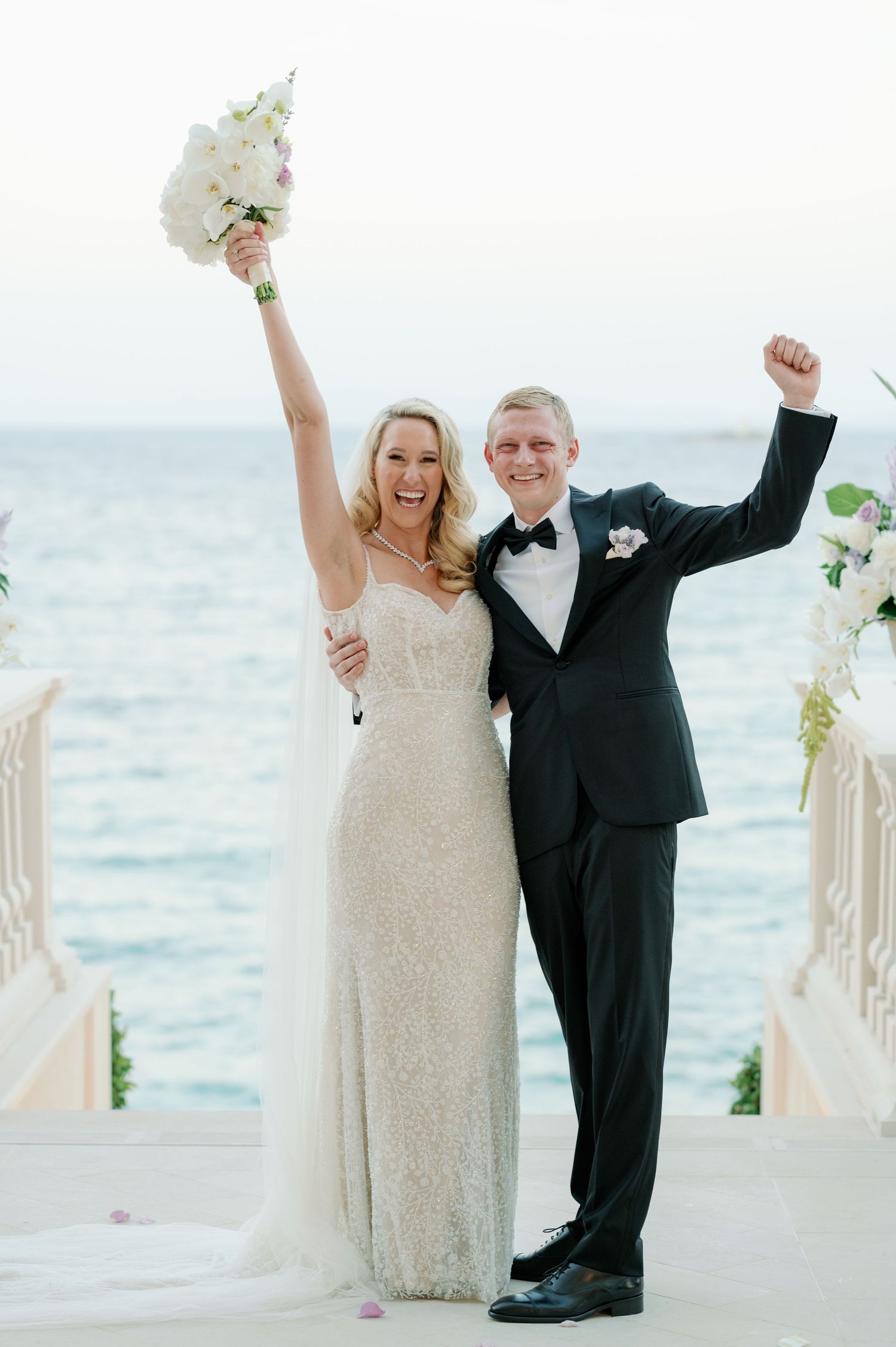 Newlyweds celebrate joyfully on their wedding day against an ocean backdrop with raised arms.