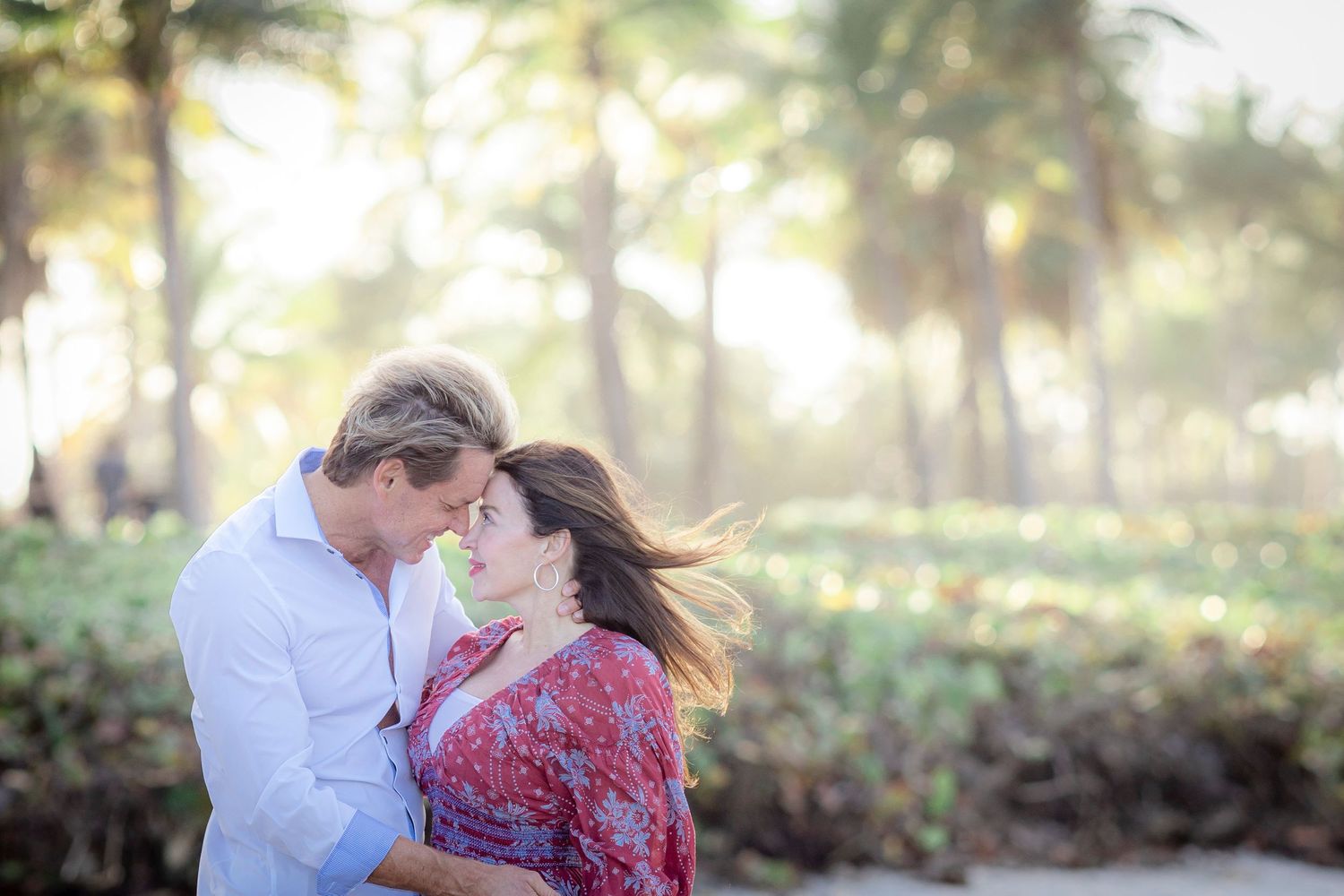 Engaged couple embracing among sunlit palm trees during an engagement session at Jupiter Beach