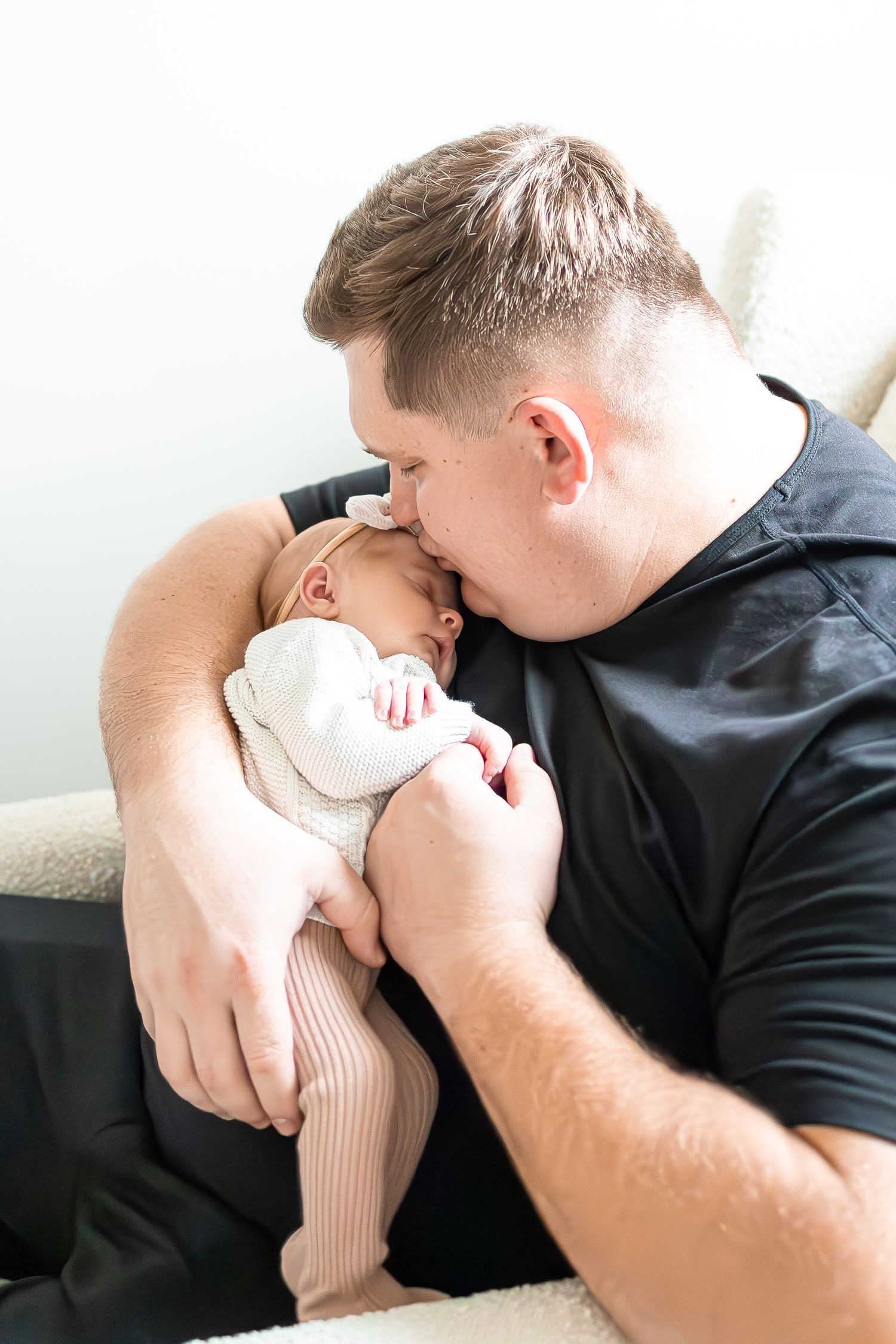 A tender moment as a caregiver in black shirt kisses a sleeping baby wearing a white bow.
