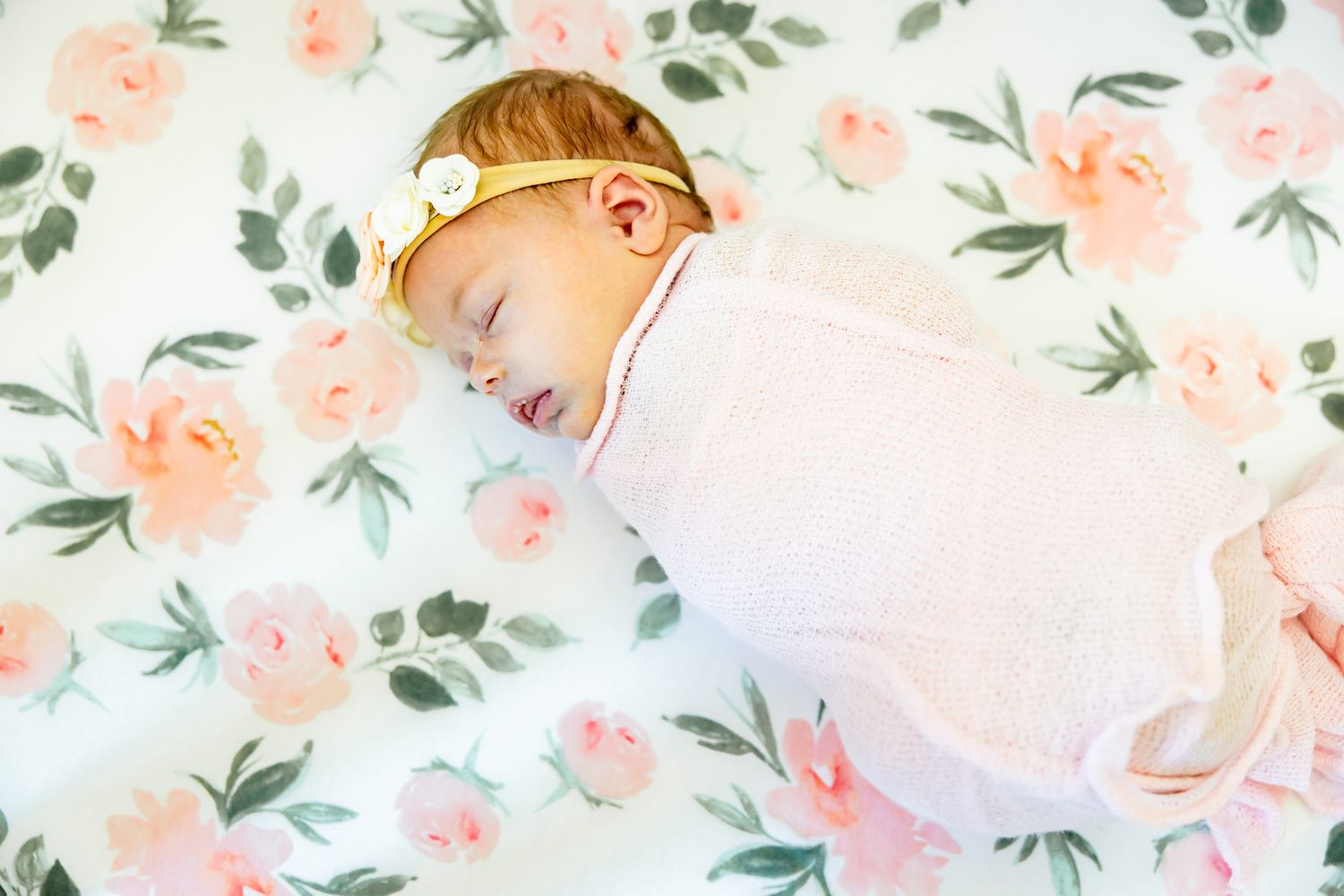 Baby girl in pink swaddle and flowery headband sleeps in her crib during newborn photos in Colorado Springs.
