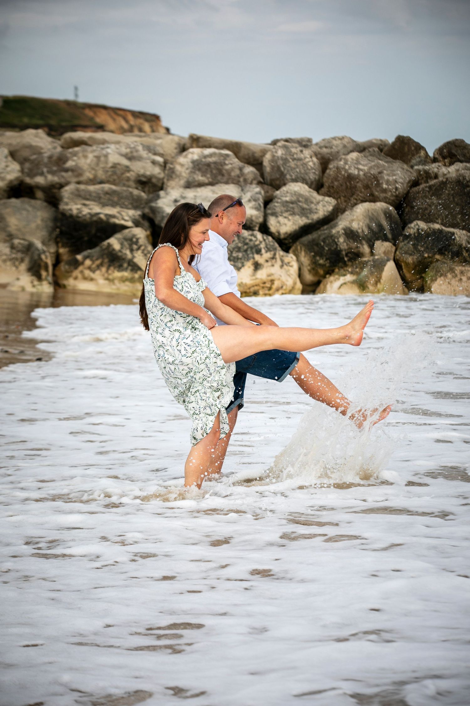 Two people splash and play in ocean waves near a rocky shoreline.
