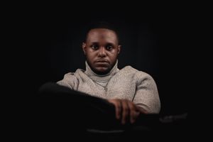 Person in beige sweater seated against black background in artistic lighting.